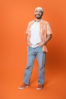 full length of happy bearded man in trendy shirt and white beanie posing with hands in pockets of blue jeans on orange background