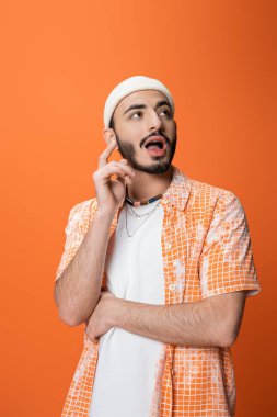 amazed bearded man in white beanie looking away and showing idea sign isolated on orange