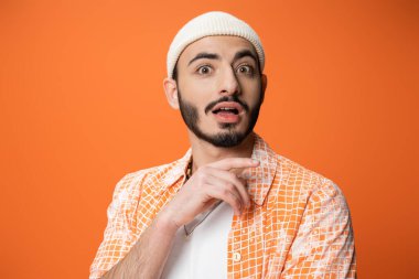 impressed bearded man in trendy shirt and white beanie looking at camera isolated on orange