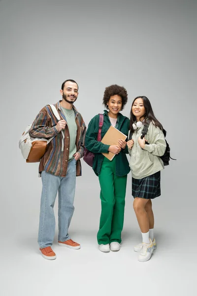 stock image full length of happy and stylish multiethnic students with backpacks and notebook standing on grey background