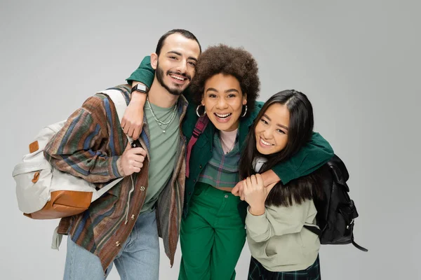excited african american woman hugging young friends with backpacks isolated on grey