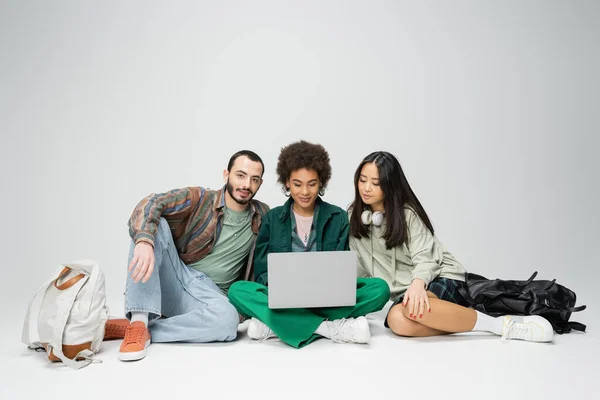 stock image full length of young african american woman sitting with laptop near stylish interracial friends on grey background