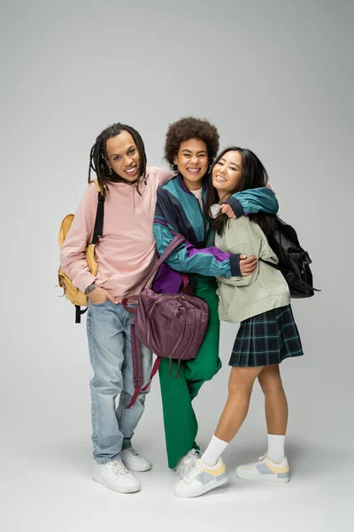 full length of joyful multicultural students with backpacks embracing while looking at camera on grey background