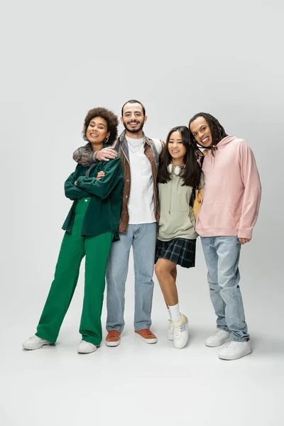 stock image full length of happy multicultural students in trendy outfit smiling at camera on grey background