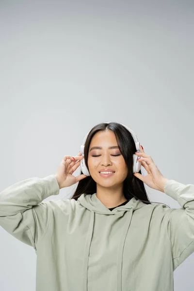 brunette asian woman in hoodie listening music in wireless headphones and smiling with closed eyes isolated on grey