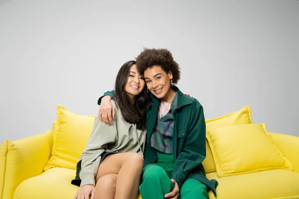 happy african american woman embracing asian friend while sitting on yellow couch isolated on grey