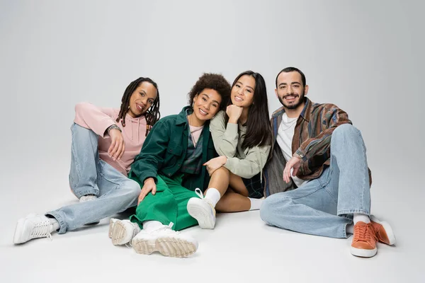 stock image full length of cheerful multicultural friends in fashionable casual attire sitting and looking at camera on grey background