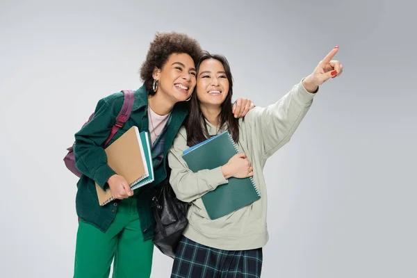 stock image smiling asian student with copybooks looking away and pointing with finger near cheerful african american friend isolated on grey