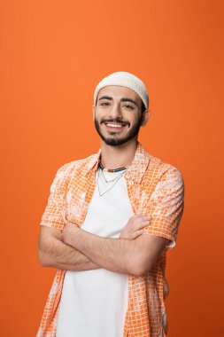 joyful bearded man in white beanie and stylish shirt standing with crossed arms isolated on orange