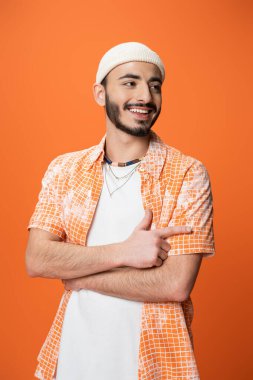 fashionable bearded man smiling and pointing with finger while looking away isolated on orange
