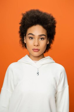 portrait of young african american woman in white hoodie looking at camera isolated on orange