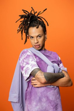 trendy multiracial man with dreadlocks posing with crossed arms isolated on orange