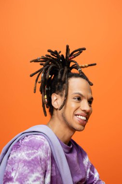portrait of positive multiracial man with dreadlocks and piercing looking away isolated on orange