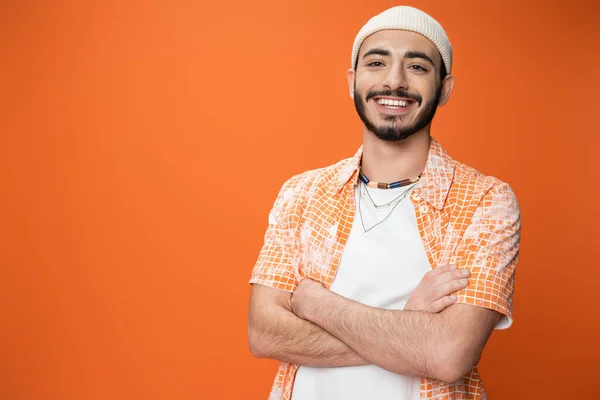 joyful bearded man in fashionable outfit posing with crossed arms isolated on orange