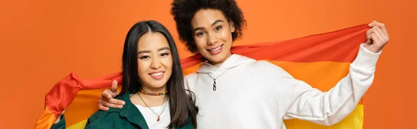 cheerful asian and african american women with lgbt flag looking at camera isolated on orange, banner