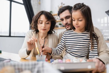Family holding paintbrushes near blurred Easter eggs in kitchen 