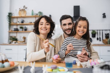 Gülümseyen aile mutfakta Easer yumurtalarını boyarken kameraya bakıyor. 