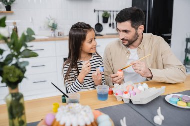 Man talking to daughter while coloring Easter eggs in kitchen  clipart
