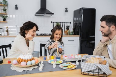 Gülümseyen kız paskalya yumurtalarını ailelerin yanında boyuyor ve mutfakta yemek yiyor. 