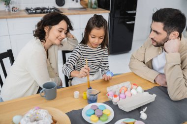 Smiling woman sitting near daughter coloring Easter eggs and husband at home  clipart