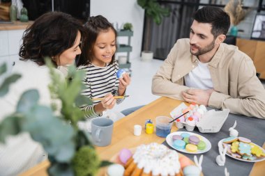 Positive parents looking at happy kid coloring Easter eggs at home  clipart