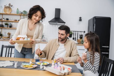 Evde aile yumurtaları boyarken elinde paskalya pastası tutan olumlu bir kadın. 