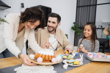 Smiling man and girl coloring Easter eggs near woman with cake at home 