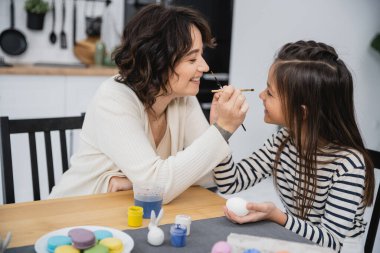 Side view of cheerful mother and child holding paintbrushes near Easter eggs and macaroons at home  clipart