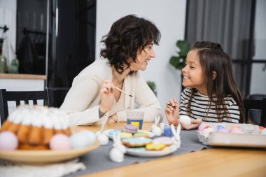 Positive mother and kid holding paintbrushes while coloring Easter eggs at home  clipart