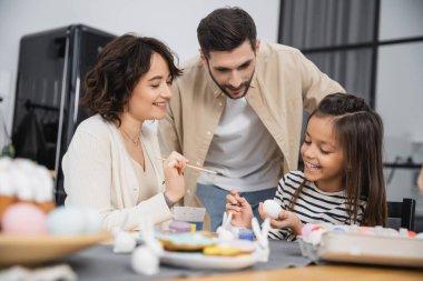 Positive daughter and mom coloring Easter eggs near father at home  clipart