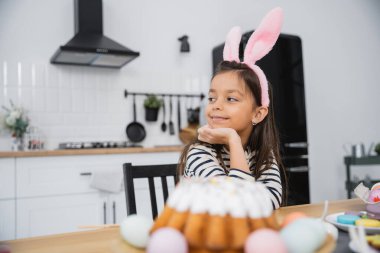 Smiling kid in headband with bunny ears near Easter cake at home 