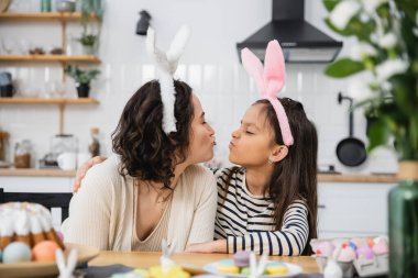 Side view of mom and daughter in headbands pouting lips near Easter food in kitchen 