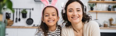 Smiling mother and child in Easter headbands looking at camera at home, banner  clipart