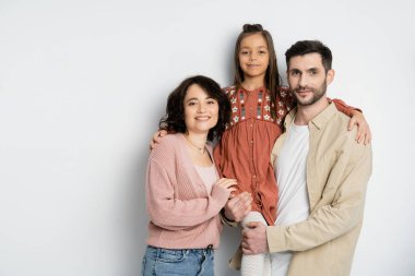 Smiling kid hugging parents and looking at camera on white background 