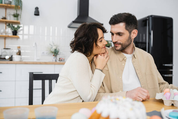 Smiling woman looking at husband near Easter eggs and cake at home 