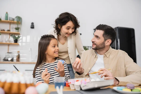 Man holding Easter egg near smiling wife and daughter in kitchen