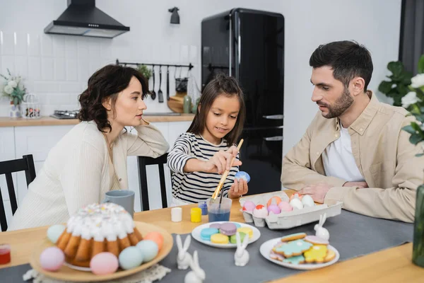 Parents talking near cheerful daughter coloring Easter eggs at home