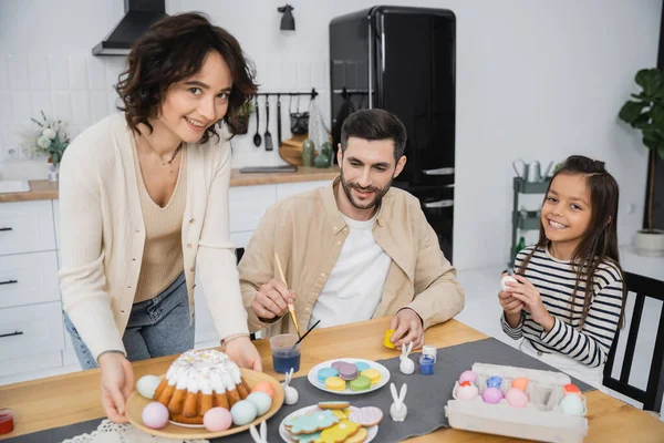 stock image Family coloring Easter eggs near cheerful mother holding plate with cake at home 