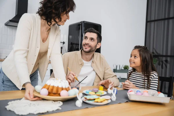 Smiling family coloring eggs near woman purring plate with Easter cake on table at home
