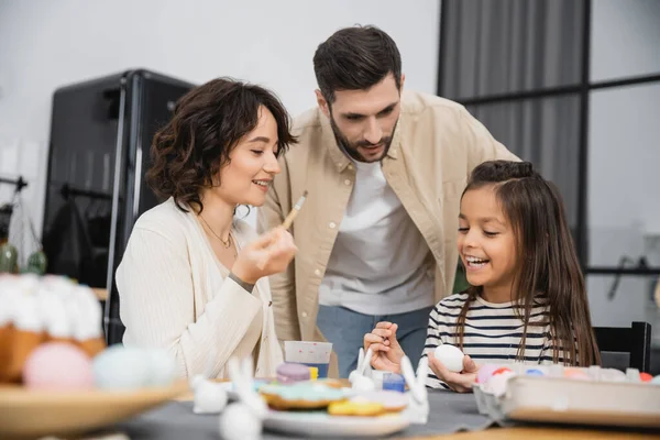 stock image Cheerful kid holding Easter egg near mother with paintbrush and dad at home 