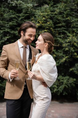 cheerful groom in suit and happy bride in wedding dress holding glasses of champagne 