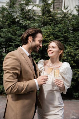 smiling groom in suit and happy bride in wedding dress clinking glasses of champagne 