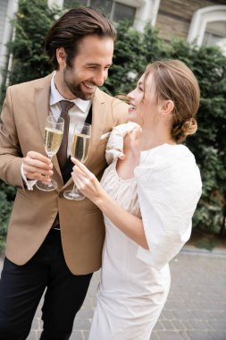 cheerful and young newlyweds clinking glasses of champagne during wedding celebration 