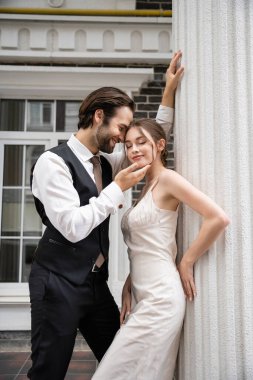 groom in formal wear smiling and touching face of pretty bride in wedding dress 