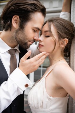 portrait of groom in formal wear touching face of pretty bride in wedding dress 