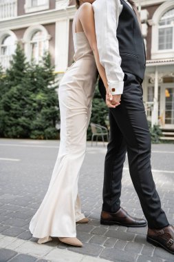 cropped view of young bride and groom in wedding dress and suit holding hands outside  clipart