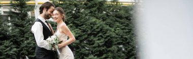 bearded groom in vest embracing cheerful bride in white dress with wedding bouquet, banner 