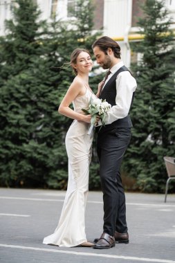 full length of bearded groom in vest embracing cheerful bride in white dress with wedding bouquet  clipart