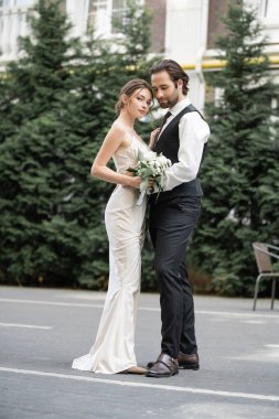 full length of bearded man in vest embracing bride in white dress with wedding bouquet 