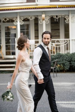 young man in suit holding hand of bride in white dress with bouquet of flowers 